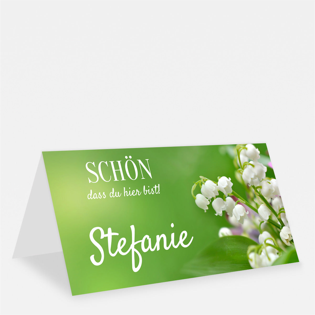 Grüne Tischkarte mit weißen Maiglöckchen, dem Namen Stefanie und dem Schriftzug „Schön, dass du hier bist!“.