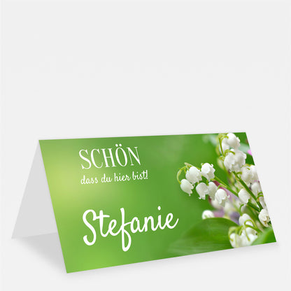 Grüne Tischkarte mit weißen Maiglöckchen, dem Namen Stefanie und dem Schriftzug „Schön, dass du hier bist!“.