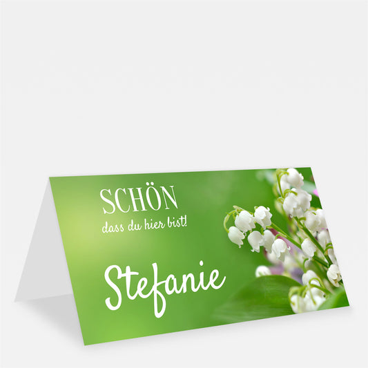 Grüne Tischkarte mit weißen Maiglöckchen, dem Namen Stefanie und dem Schriftzug „Schön, dass du hier bist!“.